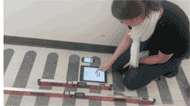 The picture displays the four instruments that were used in this study to measure ramp slope.  The four instruments are, from bottom to top, a 4 foot level, a 2 foot level, an Apple iPad, and a smaller Apple ipod Touch. The four instruments are placed nextl to each other so that the levels are parallel to the run of the ramp and the tops of the 2 Apple devices are facing toward the top of the ramp. A woman in the picture is simulating taking a measurement using the Apple iPad by touching the screen.