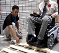 Pictures of two wheelchair users demonstrating driving with the &ldquo;Patrafour&rdquo; over  2inch obstacles-one obstacle is a step and the other obstacle  is a piece of wood