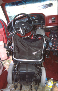 Rear view of driver&rsquo;s wheelchair in secured in docking tiedown device after the frontal crash.  The docking device is deformed such that the wheelchair is tipped slightly toward the right side of the van.  The tri-pin steering device is visible on the steering wheel.