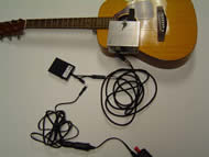 Image shows the overhead view of the entire device with connections. An AC/DC converter is just visible and is connected to a rectangular pedal through a 1/8” mono connection. The pedal is then connected to the strumming component through &frac14;” stereo connections. The strumming component is mounted to the guitar face.
