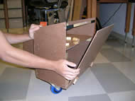 Image shows a 3/4 profile view of the functional prototype.  Image is the same as Photo 3 except it shows the flap in a more upright position, illustrating the functionality of the hinges.  Also, four metal bolts protrude through the bottom of the chamber, which is where a swivel wheel is attached.  Wooden support blocks along the side of the chamber are below the shelf.   